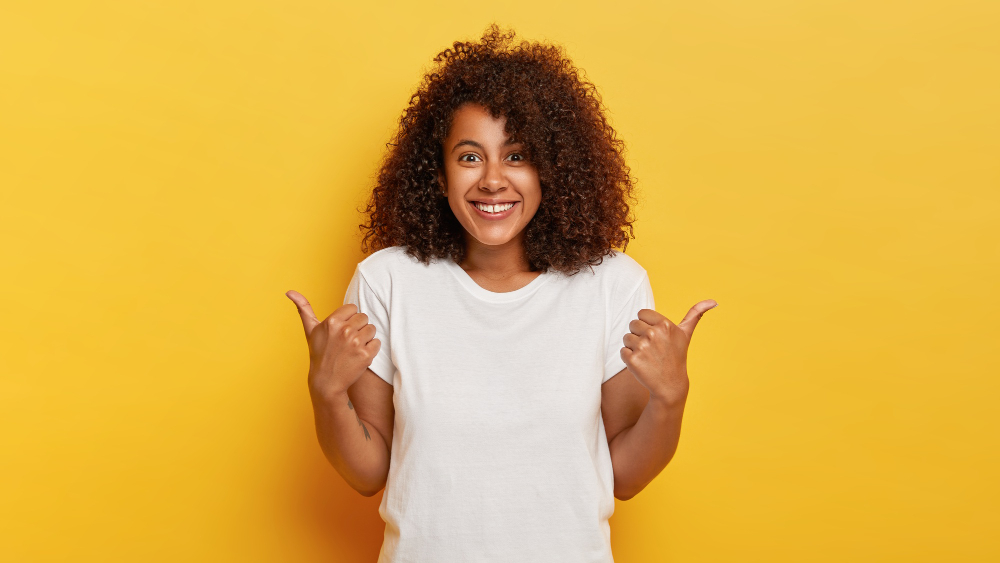 happy-curly-haired-girl-makes-thumbs-up-sign-demonstrates-support-respect-someone-smiles-pleasantly-achieves-desirable-goal-wears-white-t-shirt-isolated-yellow-wall